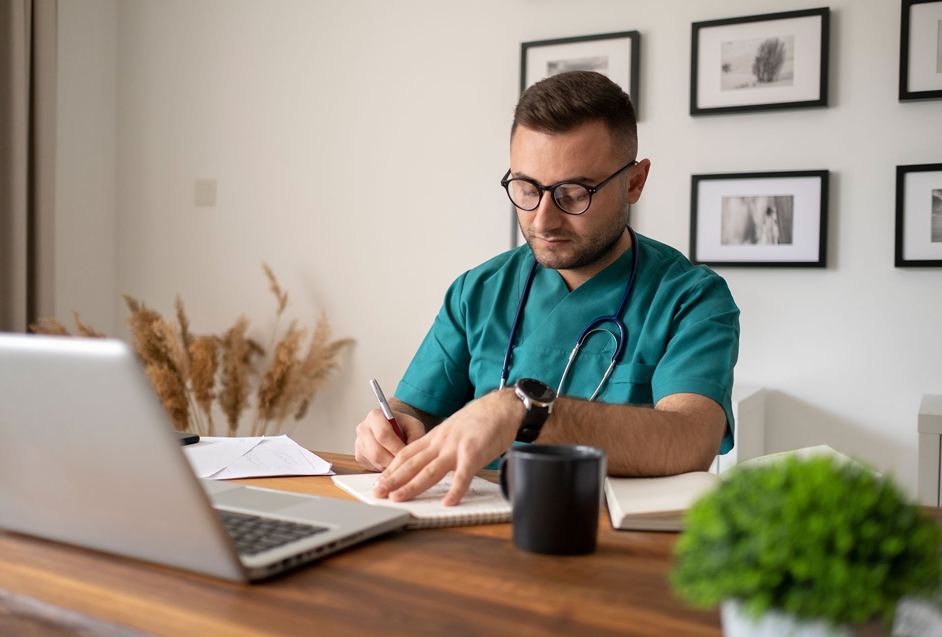 junger, lächelnder Arzt mit Stethoskop sitzt hinter Schreibtisch im Büro